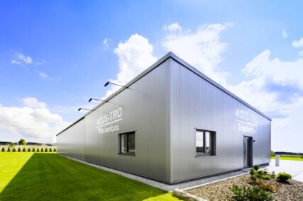 Moderne Industriehalle mit Metallfassade der Firma ACUS-TRO Trockenbau bei Tageslicht, fotografiert aus der Ecke mit Blick auf zwei Fassadenseiten. Der Himmel ist blau mit vereinzelten Wolken, das Gebäude ist von grünem Rasen und einem kleinen Kiesstreifen umgeben.