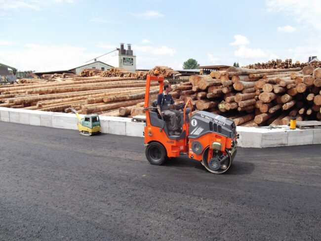 Arbeiter auf Walze bei Asphaltierungsarbeiten vor einem großen Holzlager.