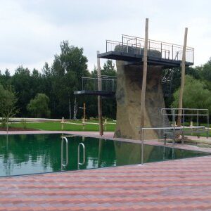 Blick auf das tiefgrüne Wasser eines Schwimmbeckens mit Zugangsstangen im Vordergrund. Der zentrale Sprungturm wirkt wie ein Felsblock mit Geländern.