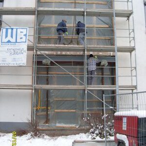Drei Bauarbeiter arbeiten auf einem eingerüsteten Fassadenbereich, der mit transparenter Folie abgedeckt ist. Der Boden ist schneebedeckt, ein Bauzaun und ein rotes Aggregat sind im Vordergrund sichtbar.