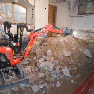 Ein roter Minibagger steht in einem Innenraum und bewegt eine große Menge an Erd- und Bauschutt. Die Wände zeigen Spuren früherer Nutzung und Sanierung.