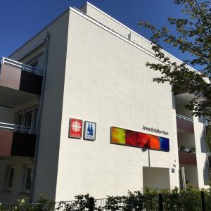 Fertiggestelltes Wohngebäude mit Balkonen und Beschilderung. An der Fassade sind das Logo der Caritas, das Kölner Stadtwappen und ein buntes Schild mit der Aufschrift „Anna-Schäffer-Haus“ zu sehen.