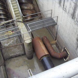 Blick in einen betonierten Schacht mit mehreren großen, schräg eingebauten Schneckenpumpen. Rostige Rohrleitungen münden in das trübe Wasser.