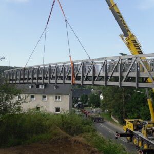 Ein Kran setzt eine Brücke aus der Luft präzise auf die vorbereiteten Fundamente – Blick von oben.