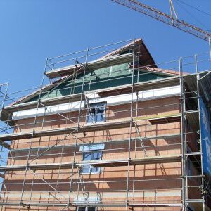 Ein mehrstöckiger Rohbau mit sichtbaren Ziegelwänden ist vollständig von einem Baugerüst umgeben. Im Hintergrund ist ein Kran zu sehen, der auf Bauarbeiten hinweist. Der Himmel ist klar und blau.