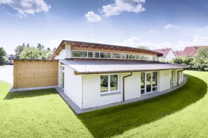 Helles Kita-Gebäude mit abgerundeter Form, Holzfassade und viel Grünfläche.