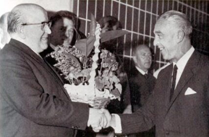 Zwei Männer im Anzug reichen sich die Hand, einer übergibt einen Blumenkorb, umgeben von weiteren Personen.
