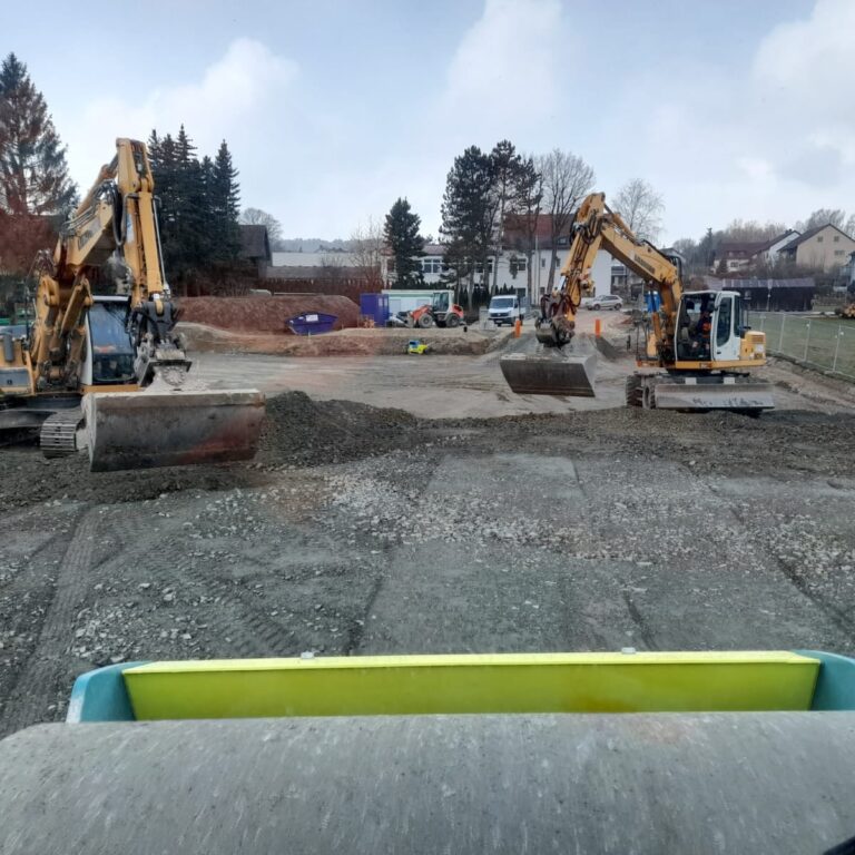 Zwei gelbe Bagger stehen sich auf einer großen Fläche gegenüber und verteilen grobes Material. Die Perspektive stammt offenbar aus einem Walzenfahrzeug.