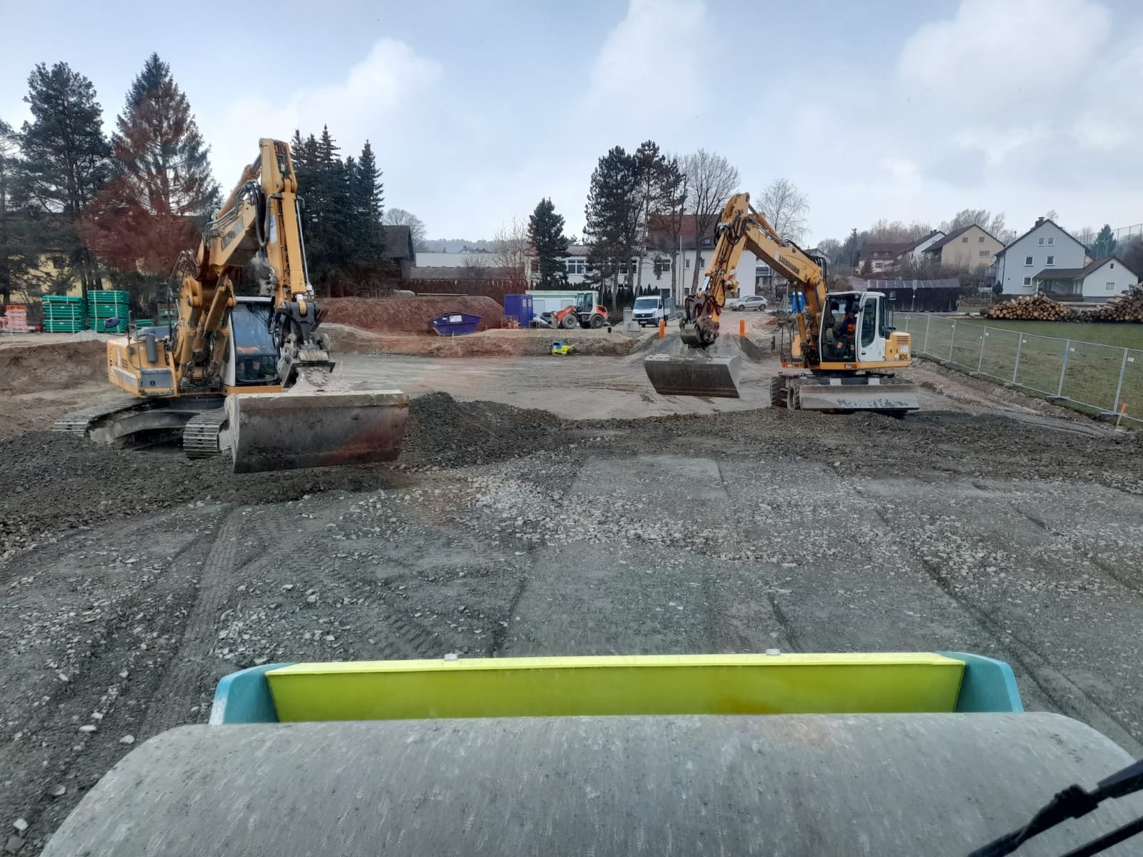 Zwei gelbe Bagger stehen sich auf einer großen Fläche gegenüber und verteilen grobes Material. Die Perspektive stammt offenbar aus einem Walzenfahrzeug.