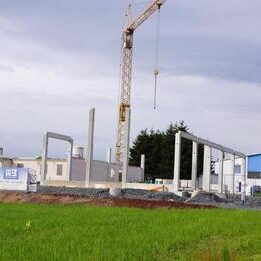 Baustelle eines Industrie- oder Gewerbebaus mit großem Kran und Rohbau-Strukturen auf freiem Feld.