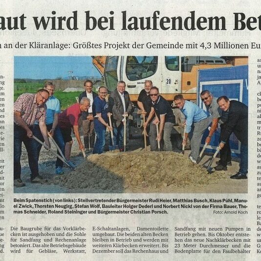 Zeitungsausschnitt mit der Überschrift „Gebaut wird bei laufendem Betrieb“ zeigt eine Gruppe von Männern mit Spaten auf einer Baustelle vor einem Bagger.