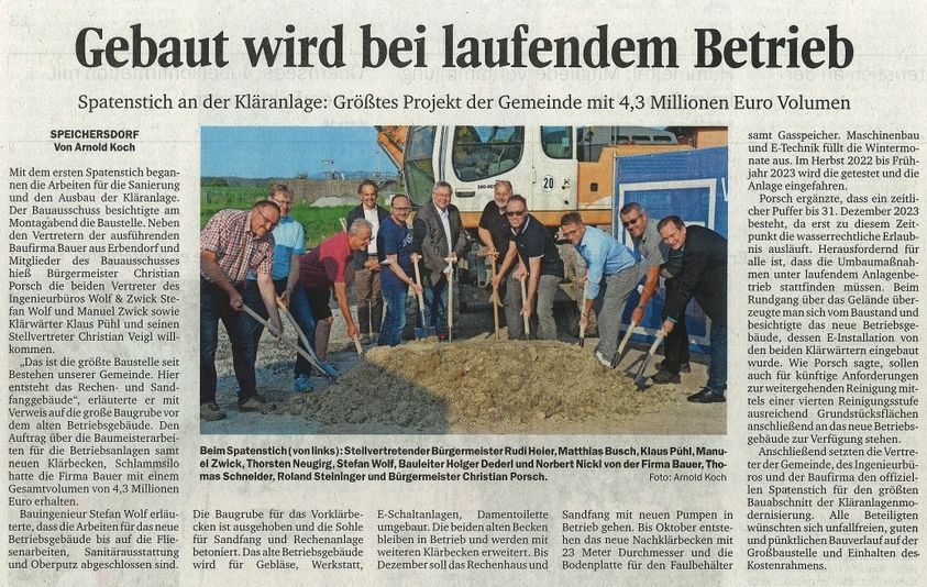 Zeitungsausschnitt mit der Überschrift „Gebaut wird bei laufendem Betrieb“ zeigt eine Gruppe von Männern mit Spaten auf einer Baustelle vor einem Bagger.