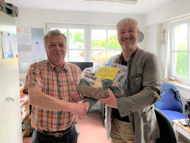 Zwei Männer lächeln und schütteln sich in einem Büro die Hand. Der rechte überreicht einen Geschenkkorb mit gelbem Zettel „Harry“.