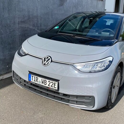 Ein silbergrauer VW ID.3 mit deutschem Kennzeichen „TIR WB 22E“ steht vor einer Betonwand auf einem asphaltierten Parkplatz.