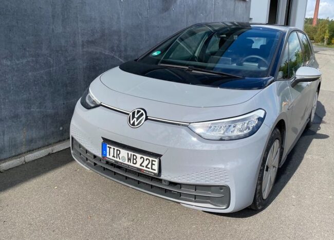 Ein silbergrauer VW ID.3 mit deutschem Kennzeichen „TIR WB 22E“ steht vor einer Betonwand auf einem asphaltierten Parkplatz.