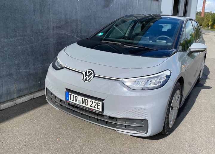 Ein silbergrauer VW ID.3 mit deutschem Kennzeichen „TIR WB 22E“ steht vor einer Betonwand auf einem asphaltierten Parkplatz.