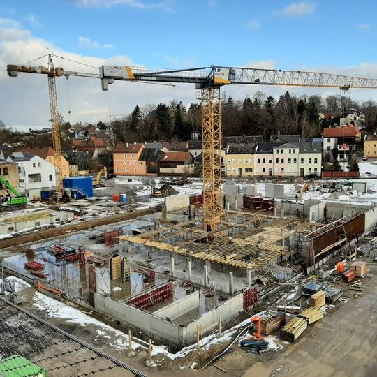 Auf einer winterlichen Baustelle entstehen mehrere Betonteile eines Gebäudes. Zwei große Kräne dominieren das Bild, im Hintergrund eine Kleinstadt.