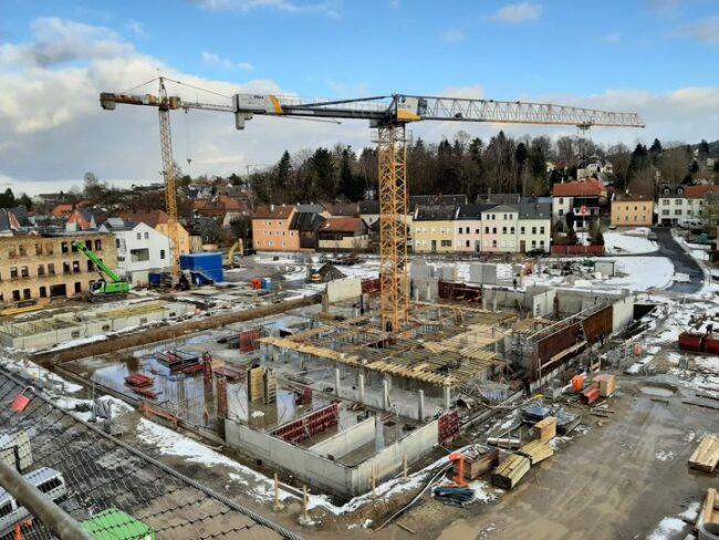 Auf einer winterlichen Baustelle entstehen mehrere Betonteile eines Gebäudes. Zwei große Kräne dominieren das Bild, im Hintergrund eine Kleinstadt.
