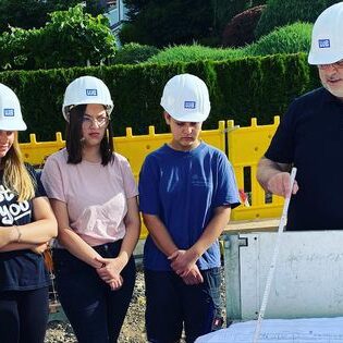 Gruppe junger Menschen mit Helmen steht an einem Bauplan und wird von einem Bauleiter unterrichtet.