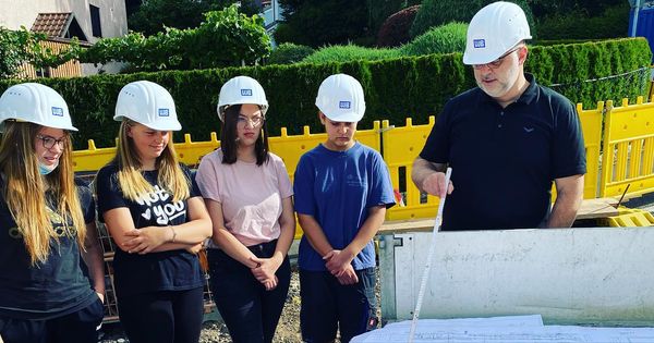 Girls Day 2022 Gruppe junger Menschen mit Helmen steht an einem Bauplan und wird von einem Bauleiter unterrichtet.