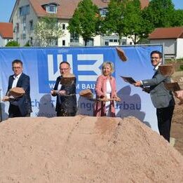 Sechs Personen werfen gemeinsam Sand in die Luft. Im Hintergrund ein Werbebanner der Firma WEB und Wohngebäude mit rotem Dach.