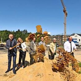 Sechs Personen werfen Sand mit Spaten in die Luft. Im Hintergrund ein altes Fabrikgebäude mit Schornstein, ein Baukran und ein Banner mit Bauprojektinformationen.
