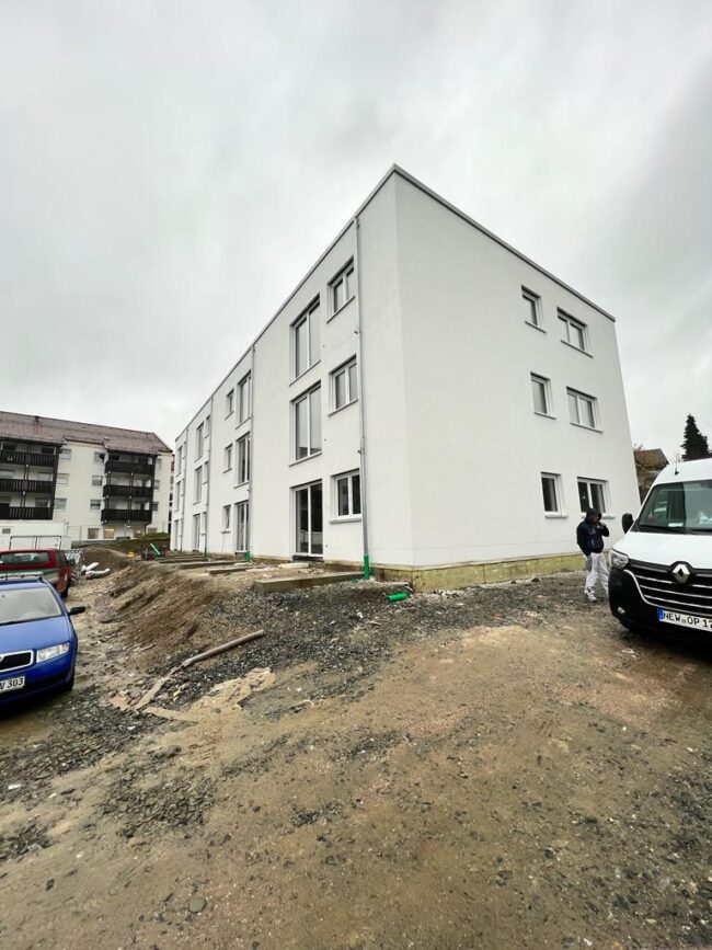 Neubau eines weißen Mehrfamilienhauses mit drei Etagen. Das Umfeld ist noch unbefestigt, es stehen mehrere Fahrzeuge und eine Person ist sichtbar.