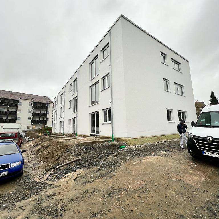 Neubau eines weißen Mehrfamilienhauses mit drei Etagen. Das Umfeld ist noch unbefestigt, es stehen mehrere Fahrzeuge und eine Person ist sichtbar.