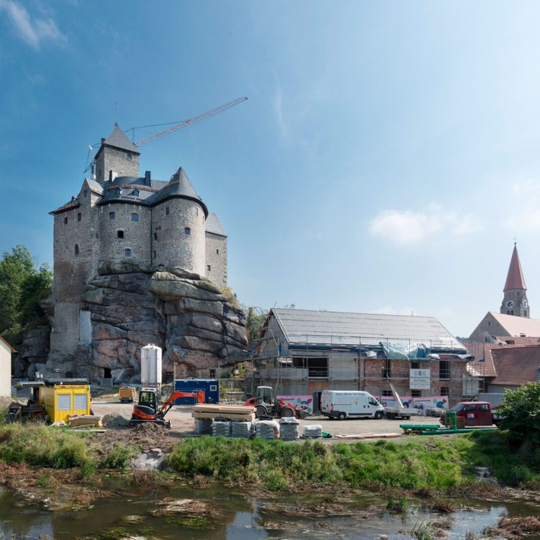 Eine massive Burganlage thront auf einem Felsmassiv. Am Fuß des Hügels laufen Bauarbeiten mit Maschinen und Materialien. Rechts eine Kirche mit Spitzdach.