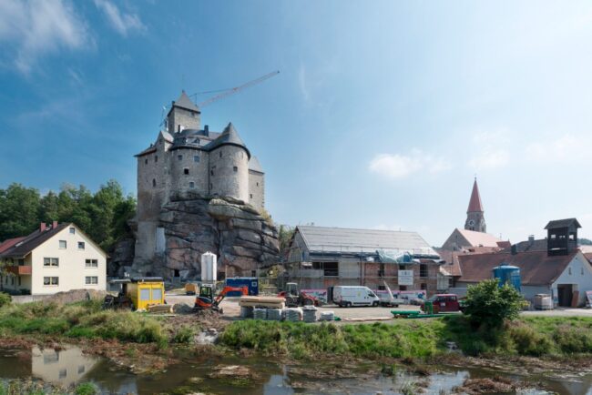 Eine massive Burganlage thront auf einem Felsmassiv. Am Fuß des Hügels laufen Bauarbeiten mit Maschinen und Materialien. Rechts eine Kirche mit Spitzdach.