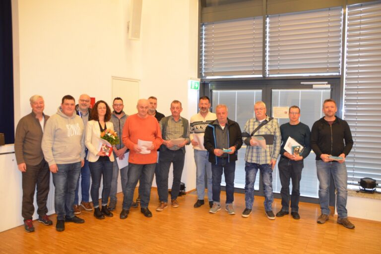 Eine Gruppe von etwa 15 Personen posiert in einem Innenraum mit Urkunden, eine Frau hält einen Blumenstrauß.