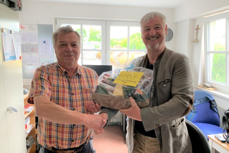 Zwei Männer reichen sich die Hand in einem Büroraum. Der rechte Mann hält einen Geschenkkorb in der Hand.