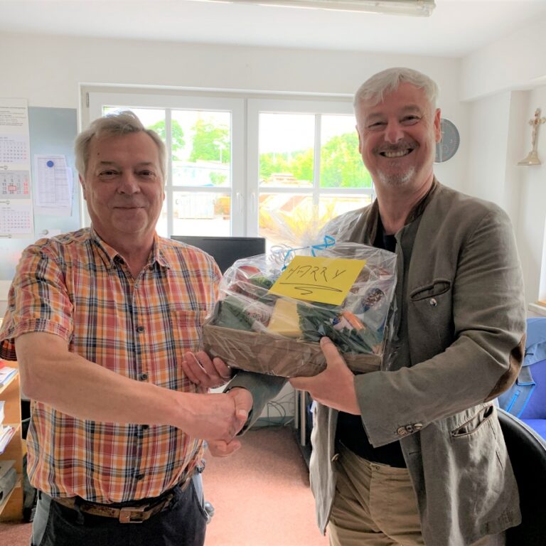 Zwei Männer reichen sich die Hand in einem Büroraum. Der rechte Mann hält einen Geschenkkorb in der Hand.